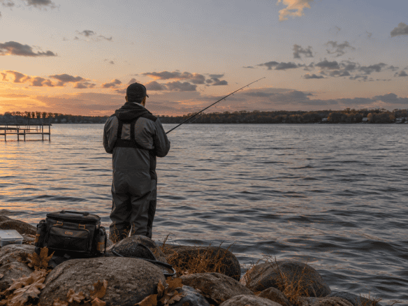 An angler enjoying Okoboji fall fishing from a rocky shoreline during a cool autumn sunset.